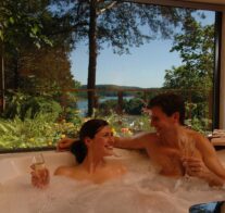 Two guests sipping champagne in a bubbling Jacuzzi with the summer view of Beaver Lake showing in the Jacuzzi-size picture window behind them.