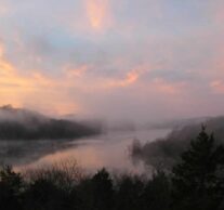 Dawn rising over a mist-shrouded Beaver Lake with multiple hues of pink, purple, blue, and the outline of the Ozark Mountains.