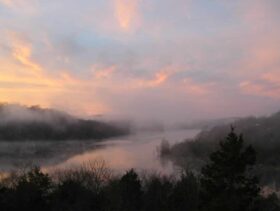 Dawn rising over a mist-shrouded Beaver Lake with multiple hues of pink, purple, blue, and the outline of the Ozark Mountains.
