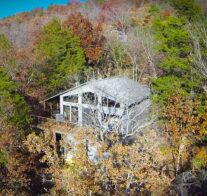 An aerial image of a cabin emerging from the autumn trees, its' lake-facing, front deck 20 feet off the ground due to the slope of the land. ground