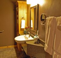 Warm light shines on the accessible lavatory, quartz counter-tops, personal mirrors, and natural pine cabinets of the bathroom.