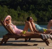 Two women sunbath on reclining lounge chairs and relax with margaritas as a pontoon boat floats along in the background.