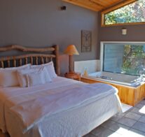 A king-size bed with luxurious linens sits next to a large, 2-person Jacuzzi and picture window looking out over Beaver Lake.