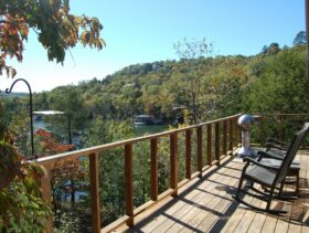 The front deck of a cabin over looking Beaver Lake on a sunny day complete with rocking chairs and a BBQ.