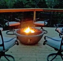 A warm flickering lava rock fire pit during twilight is surrounded by comfortable outdoor seating on the over sized lower Lake House deck with the lake and trees behind.