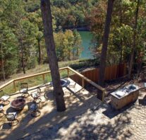 On a summer day looking from high above the expansive outdoor deck surrounded by trees has ample outdoor seating circling a lava rock fire pit as well as a large stone patio outdoor kitchen.