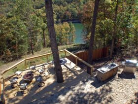 On a summer day looking from high above the expansive outdoor deck surrounded by trees has ample outdoor seating circling a lava rock fire pit as well as a large stone patio outdoor kitchen.