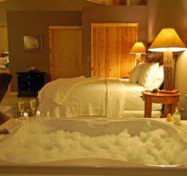A night-time scene with a bubble-filled Jacuzzi with two champagne glasses awaits in the foreground with the king-size bed and nightstands behind.